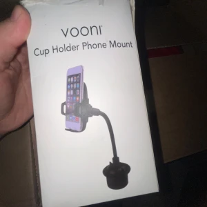 Vooni Cup Holder Phone Mount - Säljer en praktisk telefonhållare från Vooni som passar i bilens mugghållare. Den har en flexibel arm för enkel justering och säker montering av din telefon. Perfekt för handsfree-användning under bilresor. Kompakt och lätt att installera.