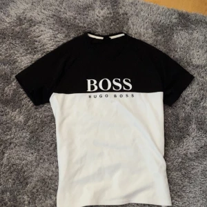 Svart och vit t-shirt från Hugo Boss - Snygg svart och vit t-shirt från Hugo Boss med stor logga på bröstet. Perfekt för en stilren och modern look. Kortärmad och tillverkad i mjukt material för extra komfort.