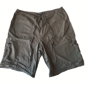 Gröna shorts från Angels - Säljer ett par gröna shorts från Angels med fickor och dekorativa ringdetaljer på sidorna som kan användas för att justera längden. Tror att storleken är m/l.
