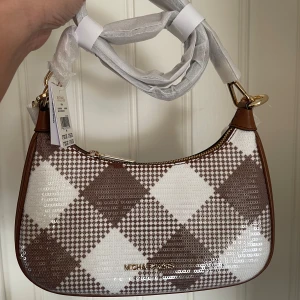 Rutig axelväska från Michael Kors - Snygg axelväska från Michael Kors i ett rutigt mönster med bruna och vita nyanser. Väskan har en dragkedja upptill och är perfekt för att bära dina nödvändigheter med stil. Den har också en justerbar axelrem för bekvämlighet. Inget kvitto då denna är köp via vinted från en som har god omdöme. 