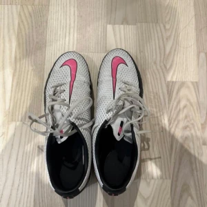 Nike phantom gt fotbollsskor  - Snygga vita fotbollsskor från Nike med rosa detaljer och svart sula. Skorna har snörning och är perfekta för inomhusfotboll. Den rosa Nike-loggan ger en cool kontrast mot det vita.