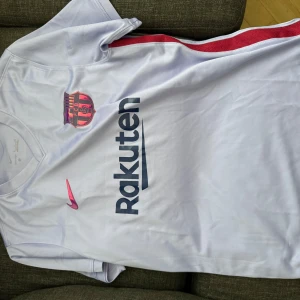  Lilla Barca tröja  - Säljer en vit fotbollströja från Nike med klubbmärke på bröstet och sponsorloggor. Tröjan har korta ärmar och en röd rand längs sidan. Perfekt för fotbollsfans!