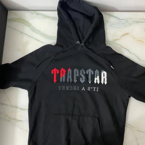 Svart hoodie från Trapstar - Säljer en svart hoodie från Trapstar med tryck i rött och vitt på framsidan. Den har en klassisk design med dragsko i huvan och en stor ficka framtill. Perfekt för en avslappnad stil.
