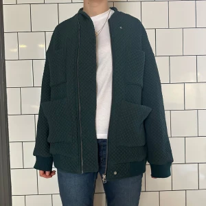 Mörkgrön quiltad bomberjacka - Säljer en snygg mörkgrön quiltad bomberjacka från Carin Wester med dragkedja framtill. Jackan har en oversized passform och långa ärmar med ribbade muddar. Perfekt för en cool och avslappnad stil.
