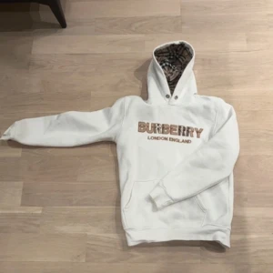 Vit hoodie från Burberry - Säljer en stilren vit hoodie från Burberry med logotypen broderad i brunt på framsidan. Hoodien har en klassisk känguruficka och en fodrad huva med Burberrys ikoniska rutmönster. Perfekt för en avslappnad och trendig look. Tar även emot byten.