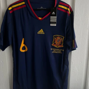 Blå fotbollströja från Adidas - Säljer en blå fotbollströja från Adidas med korta ärmar. Tröjan har gula och röda detaljer samt ett emblem på bröstet. Nummer 6 och namnet A. Iniesta är tryckt på baksidan. Perfekt för fotbollsfans!