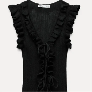 Svart topp från Zara - Snygg svart topp från Zara med volangdetaljer och v-ringning. Toppen har en knytning framtill och är ärmlös, vilket ger en elegant look. Perfekt för olika tillfällen!