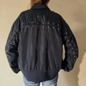 Mörkblå bomberjacka med snörning - Snygg mörkblå bomberjacka med unik snörningsdetalj på ryggen och ärmarna. Perfekt för en cool och avslappnad stil. Bilderna är lånade men skriv för egna!! 