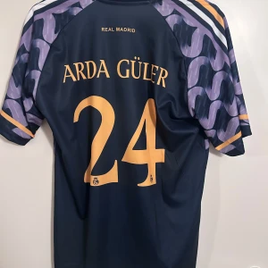Real Madrid fotbollströja med Arda Güler 24 - Snygg Real Madrid fotbollströja från Adidas med Arda Güler och nummer 24 på ryggen. Tröjan har ett unikt mönster i lila och svart med korta ärmar. Perfekt för alla fans av laget!