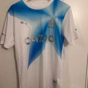 Marseille FC tröja - Säljer en snygg vit fotbollströja från Puma med blått mönster på framsidan. Tröjan har korta ärmar och en rund halsringning. Perfekt för fotbollsfans som vill visa sitt stöd med stil.