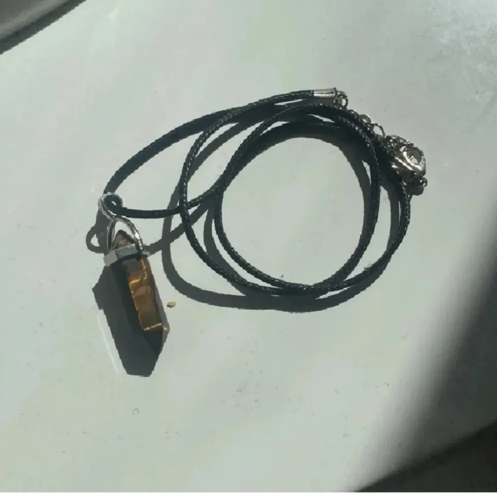 Snyggt halsband med en tigeröga-pendel i en silverfärgad infattning. Hängsmycket har en unik, naturlig mönstring i brunt och guld. Kedjan är svart och justerbar med ett klolås. Perfekt för att ge en bohemisk touch till din stil.. Asusteet.