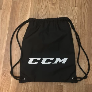 Svart gympapåse från CCM - Säljer en svart gympapåse från CCM med vit logga och snörning. Perfekt för att bära med sig träningskläder eller andra småsaker. Enkel och stilren design med plats för namn på etiketten. Helt oanvänd 