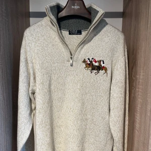 Beige stickad tröja från Polo by Ralph Lauren - Säljer en stilren beige stickad tröja från Polo by Ralph Lauren med en broderad polospelare på bröstet. Tröjan har en dragkedja vid halsen och långa ärmar. 