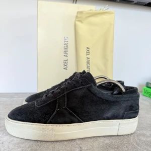 Svarta sneakers från Axel Arigato - Snygga svarta sneakers från Axel Arigato i mocka med vit sula. Skorna har en stilren design med snörning och är perfekta för en avslappnad look. De kommer med originalförpackning och påse.