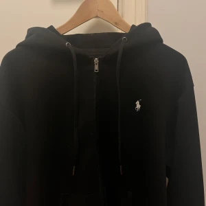 Svart hoodie med dragkedja från Ralph Lauren - Säljer en stilren svart hoodie från Ralph Lauren med dragkedja och justerbar huva. Den har en liten broderad logga på bröstet och praktiska fickor framtill. Perfekt för en avslappnad look. Tröjan är i fint skick och ja säljer den då den är lite för stor!
