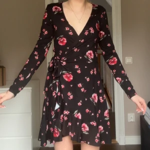 Svart blommig omlottklänning - Snygg svart omlottklänning med röda och rosa blommor. Klänningen har en v-ringad hals och långa ärmar, perfekt för både vår och höst. Den knyts i midjan för en smickrande passform.
