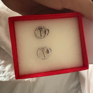 Guess örhängen i silver med stenar - Snygga örhängen från Guess i silverfärgad metall. De är runda med glittrande stenar runt hela cirkeln och har en liten nyckel som detalj inuti. Perfekta för att ge din look lite extra bling!