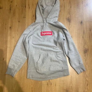 Grå Supreme hoodie med röd boxlogo - Supreme hoodie i ljusgrått med den klassiska röda boxlogon på bröstet. Hoodien är köpt någon gång mellan 2013–2015 i New York av en familjevän, så exakt modellnamn är oklart. Tröjan har huva med snörning, känguruficka och ribbade muddar. Tillverkad i mjuk bomull – perfekt för chill dagar. Supreme-tag på sidan och insidan av kragen. Äktheten är garanterad.
