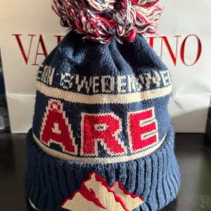 Blå Åre mössa med tofs - Säljer en stickad blå mössa från Åre med stor tofs i blått, vitt och rött. Mössan har ribbad kant, broderad bergsdetalj och texten 'ÅRE' i rött framtill. Perfekt för kalla dagar och vintersäsongen.