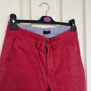 Röda slim fit chinos från Dressmann - Snygga röda chinos från Dressmann i slim fit-modell. Byxorna har klassiska fickor fram och bak, bälteshällor och stängs med dragkedja och knapp. Tillverkade i bomull med lite stretch för extra komfort. Finns en liten fläck baktill (se bild)
