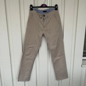 Beige chinos slim fit från Dressmann - Snygga beige chinos från Dressmann i slim fit-modell. Byxorna har klassiska sidfickor, två bakfickor med knapp och bälteshällor. Tillverkade i 98% bomull och 2% elastan för en skön stretchig känsla. Perfekta för en clean och stilren look. Har några små fläckar på byxan. 