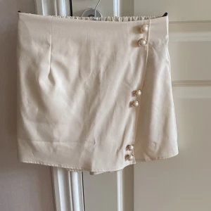 Beige/krämvit kortkjol med pärlknappar - Superfin kjol med snygga pärlknappar längs sidan och elastisk midja bak. Kjolen har en stilren och enkel design som känns både trendig och klassisk. Perfekt att matcha med en crop top eller stickad tröja för en cool look. Shorts baktill. Aldrig använda 