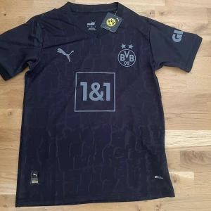 Svart Borussia Dortmund fotbollströja Puma - Snygg svart Borussia Dortmund fotbollströja från Puma med subtilt mönster och gråa detaljer. Kortärmad med BVB-logga och sponsortryck på bröstet. Tillverkad i lätt och ventilerande DryCell-material som passar perfekt för träning eller match.