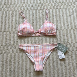 Rutig bikini i pastell från H&M - Superfin bikini från H&M i pastelliga toner med rutigt mönster i rosa, orange och vitt. Överdelen har smala axelband och en söt knut framtill. Tillverkad i mjuk polyesterblandning som torkar snabbt – perfekt för stranden! OANVÄND.                               Överdel - storlek 38, Underdel - storlek 34‼️