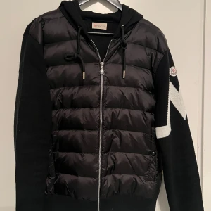 Moncler Cardigan - Snygg svart hoodie från Moncler med vadderad front i polyester och stickade ärmar. Stora vita zigzag-detaljer på ena ärmen och klassisk Moncler-logga på axeln. Huva med snörning och hel dragkedja framtill. Perfekt för dig som gillar streetstyle. Passar M och S , skriv för mer info , pris kan diskuteras 