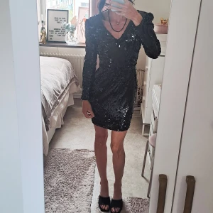 Svart glittrig paljettklänning - Säljer en svart kort klänning med glittriga paljetter och v-ringad hals. Klänningen har lång ärm och markerade axlar som ger en cool siluett. Perfekt för dig som vill sticka ut med en festlig look. Sitter snyggt och har en tight passform. Skönt innedag som gör att paljetter inte "kliar". 