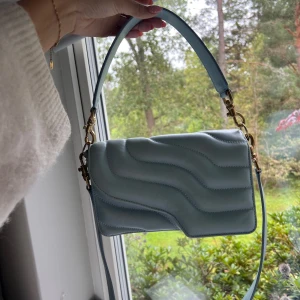 ATP väska  - Superfin ljusblå axelväska från ATP Atelier i mjukt skinn. Väskan har vågformad quiltad design, guldiga metalldetaljer och både kort och lång rem. Perfekt storlek för det viktigaste och ger en trendig touch till din outfit. Finns en defekt på det kortare bandet, men inget som märks av 