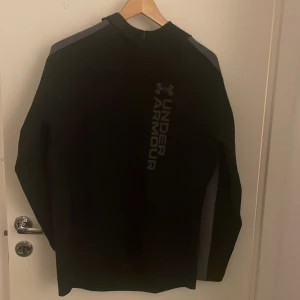 Svart hoodie från Under Armour S - Svart hoodie från Under Armour med grå detaljer längs axlar och ärmar. Tröjan har en snygg logga framtill och reflekterande detaljer på ryggen. Perfekt för träning eller chill, med lång ärm och huva. Materialet är mjukt och stretchigt syntet.
