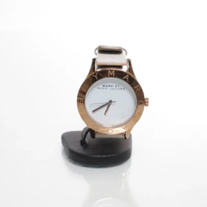 Marc Jacobs klocka med vit läderrem - Stilren klocka från Marc Jacobs med vit urtavla och roséguldfärgat runt boetten. Klockan har en vit läderrem och minimalistisk design med tre visare. Runt boetten står MARC BY MARC JACOBS ingraverat. Perfekt accessoar för en clean look.                                        Behöver att nytt batteri.