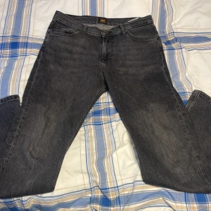 Svarta Lee jeans straight fit - Säljer ett par svarta jeans från Lee med klassisk femficksdesign och raka ben. Jeansen har en snygg tvättad look och Lee-logga på knappen och bakfickan. Perfekta för dig som gillar en enkel och tidlös stil.