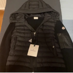 Svart Moncler pufferjacka med huva - Svart pufferjacka från Moncler med stickade ärmar och quiltad front. Jackan har huva, dragkedja framtill och flera fickor, inklusive en på ärmen med Moncler-logga. Perfekt för dig som vill ha en snygg och sportig look med premiumkänsla.
