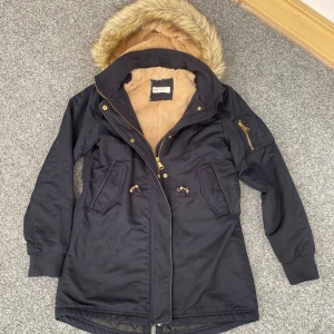 Marinblå jacka med fuskpäls - Mörkblå jacka/kappa från H&M med mysig beige fuskpäls i huva och överkropp, quiltat foder nedtill och dragkedja med knappar. Jackan har huva med fuskpäls, snörning i midjan och flera fickor, perfekt för kalla dagar!🙌🏻Luvan är avtagbar också🤩Storlek 146🩷Använd några gånger! Hör av dig❤️