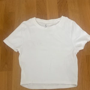 Vit croppad t-shirt i ribbad bomull - Enkel och fräsch vit t-shirt med korta ärmar och croppad passform. Tillverkad i ribbad bomull som ger en snygg struktur. Perfekt till jeans eller kjol för en clean look.