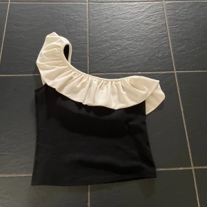 Svart one shoulder topp med volang - Snygg svart topp med en axel och stor vit volang längs halsringningen. Perfekt för dig som vill sticka ut med en cool och trendig look. Toppen är figurnära och har en unik design med asymmetrisk axel.