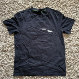 Svart BOSS t-shirt med tryck - Svart t-shirt från BOSS med coolt grafiskt tryck framtill och detaljerat mönster med små silvriga nitar på ryggen. Klassisk rund hals och korta ärmar. Tillverkad i mjuk bomull som känns skön mot huden. Perfekt för dig som gillar stilrena och moderna plagg.