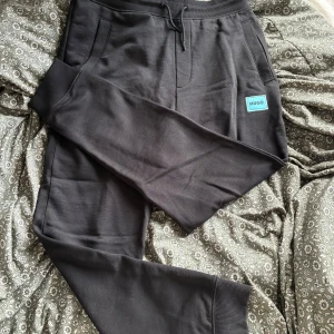 Svarta mjukisbyxor från Hugo och t shirt  - Svarta mjukisbyxor från Hugo med ribbade muddar, snörning i midjan och en blå Hugo-logga på vänster ben. Byxorna har en relaxed fit och är tillverkade i mjuk bomull för maximal komfort. Perfekta för chill dagar eller när du vill ha en clean streetstyle.T shirt är med också.