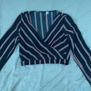 Randig omlottopp från H&M Divided - Säljer en svart omlottopp från H&M Divided med röda och vita ränder. Toppen är croppad, har lång ärm och v-ringning. Perfekt för dig som gillar trendiga och bekväma plagg med snygg passform.