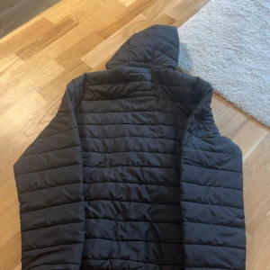 Svart pufferjacka med huva - Säljer en svart pufferjacka med huva och dragkedja framtill. Jackan har ett quiltat mönster med horisontella sömmar och är perfekt för kyligare dagar. Enkel och stilren design som funkar till det mesta. Passar dig som gillar en clean look.