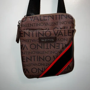 Axelväska från Valentino - Snygg brun axelväska från Valentino med logomönster över hela väskan. Justerbar svart axelrem och flera dragkedjefack. Perfekt storlek för mobilen och det viktigaste. Går ej att få tag på längre, ingen liknande på annons.