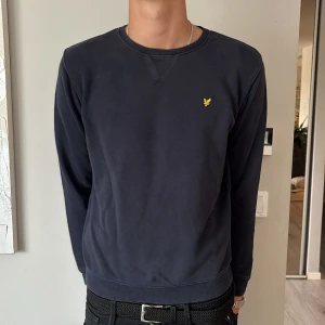 Marinblå sweatshirt från Lyle & Scott - Marinblå sweatshirt från Lyle & Scott med klassisk rund hals och gul broderad logga på bröstet. Tröjan har långa ärmar och är tillverkad i mjuk bomull, perfekt för en chill och stilren look.
