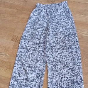 Leopardmönstrade wide byxor från H&M - Säljer ett par vita byxor från H&M med svart och blått leopardmönster. Byxorna har bred passform, resår i midjan och är tillverkade i ett lätt och luftigt material som passar perfekt för varmare dagar.