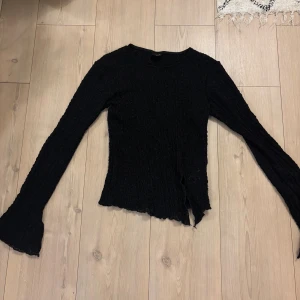 Svart långärmad ribbad topp med slits  - Svart långärmad topp med ribbad struktur och lätt utsvängda ärmslut. Tunt material som ger en cool och edgy vibe. Perfekt att styla med jeans eller kjol för en trendig look.