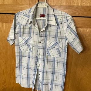 Levis Rutig kortärmad skjorta - Snygg kortärmad skjorta från Levis med rutigt mönster i vitt, blått och grönt. Skjortan har två bröstfickor med lock och tryckknappar samt klassisk krage. Perfekt för dig som gillar retrovibbar och vill sticka ut lite extra.