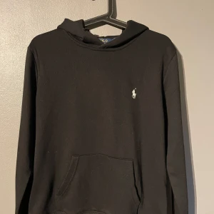 Svart hoodie från Polo Ralph Lauren - Svart hoodie från Polo Ralph Lauren med klassisk vit broderad logga på bröstet. Tröjan har huva, känguruficka och långa ärmar. Tillverkad i mjuk bomull, perfekt för chill dagar eller när du vill ha en clean look. Ops! Storlek m men passar S