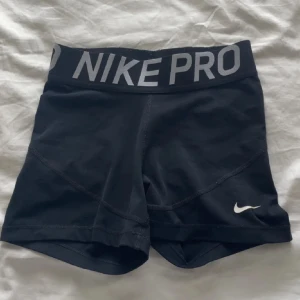 Nike Pro svarta träningstights-shorts - Svarta Nike Pro shorts med bred elastisk midja och stor logga i grått. Tight passform och korta ben, perfekta för gymmet eller sport. Materialet är mjukt och stretchigt syntet som andas bra. Klassisk Swoosh-detalj på benet.