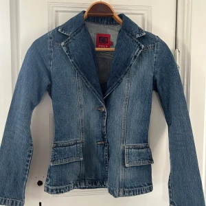 Jeanskavaj från ONLY med knappar - Säljer en snygg jeanskavaj från ONLY i klassisk blå denim. Jackan har två knappar framtill, breda slag och två fickor med lock. Långärmad och figursydd modell som ger en cool och tidlös look. 